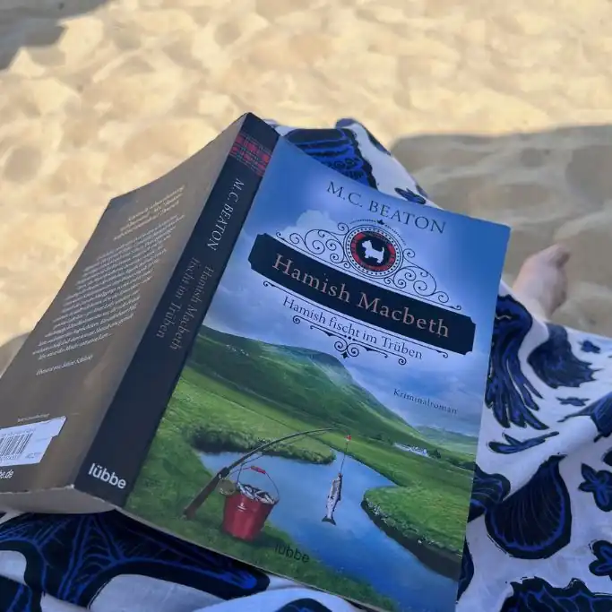 ine Person liegt am Sandstrand und hält den Kriminalroman „Hamish Macbeth – Hamish fischt im Trüben“ von M.C. Beaton geöffnet in der Hand. Auf dem Cover ist eine grüne schottische Landschaft mit einem Fluss und einer Angel zu sehen.