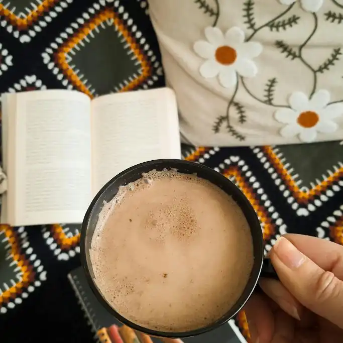Vogelperspektive auf eine Hand, die eine dunkle Tasse mit schäumender heißer Schokolade hält. Im Hintergrund liegen ein aufgeschlagenes Buch, eine gemusterte Decke und ein Kissen mit Blümchenmuster.