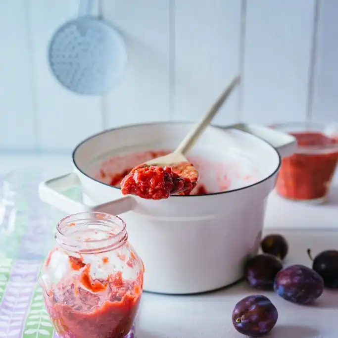 in weißer Kochtopf mit Pflaumenmus, aus dem ein Holzlöffel eine Portion rot-violette Marmelade hochhält. Im Vordergrund stehen ein offenes Einmachglas und einige frische, dunkle Pflaumen.