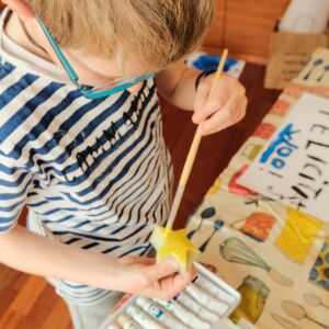 Draufsicht auf ein Kind mit Brille und blau-weiß gestreiftem Shirt, das am Basteltisch sitzt. Es hält einen Holzstern in der Hand und bemalt ihn aufmerksam mit gelber Farbe. Auf dem Tisch liegen Farbtuben und bemaltes Papier.