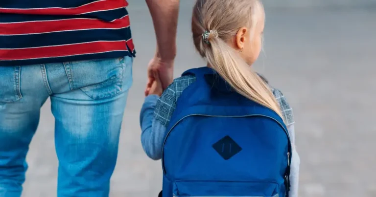 Querformatiges Motiv von hinten fotografiert: Ein blondes Mädchen mit Pferdeschwanz und blauem Rucksack geht an der Hand eines Erwachsenen im gestreiften Shirt und Jeans auf dem Weg zur Schule.