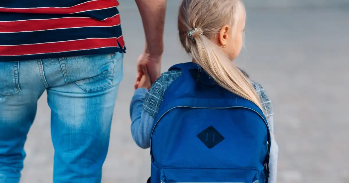 Querformatiges Motiv von hinten fotografiert: Ein blondes Mädchen mit Pferdeschwanz und blauem Rucksack geht an der Hand eines Erwachsenen im gestreiften Shirt und Jeans auf dem Weg zur Schule.