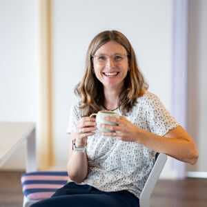 Sympathisches Portrait einer Frau mit halblangen braunen Haaren und Brille. Sie trägt ein helles, fein gemustertes Oberteil, sitzt leicht seitlich auf einem Stuhl und hält lächelnd eine helle Keramiktasse vor sich. Der Hintergrund ist hell und freundlich gestaltet.