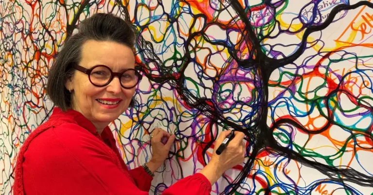 Marina Linder zeichnet lächelnd mit einem schwarzen Stift an einem großflächigen, bunten Neurografik-Kunstwerk.