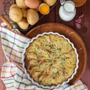 Frisch gebackener Kartoffelauflauf in einer weißen Rilleneinflorm auf einem Holzuntersetzer.