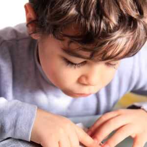 Porträt-Nahaufnahme eines kleinen Jungen mit braunen Locken im grauen Pullover. Er schaut mit gesenktem Blick und voller Fokus nach unten auf eine Buchseite, während seine Finger die Zeilen verfolgen.