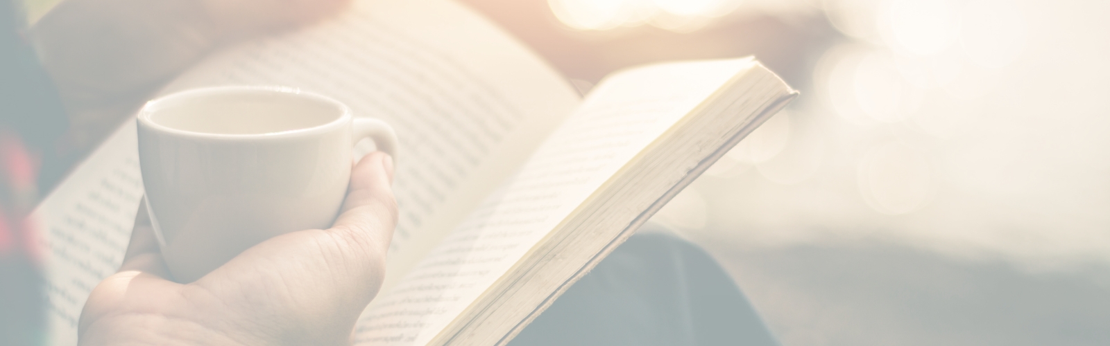 Sanft ausgeleuchtetes, helles Banner im Querformat. Eine Person hält eine weiße Tasse Kaffee oder Tee in der Hand und liest in einem aufgeschlagenen Buch, im Hintergrund ist weiches, glitzerndes Sonnenlicht zu sehen.