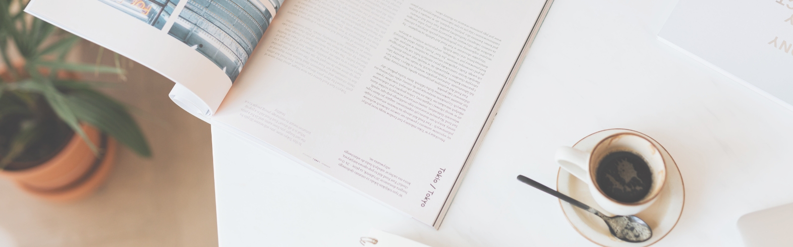 Aufgeschlagenes Lifestyle-Magazin neben einer Tasse Kaffee und einer Zimmerpflanze auf einem hellen Tisch in Vogelperspektive.