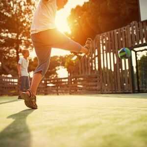 Dynamische Aufnahme in Bodenperspektive auf einem Schulhof oder Sportplatz: Ein Kind tritt im Gegenlicht der Abendsonne nach einem grün-blauen Ball. Im unscharfen Hintergrund ist ein zweites Kind zu sehen.
