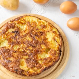 Frisch gebackene klassische spanische Tortilla auf einem runden Holzbrett mit Kartoffeln und Eiern im Hintergrund.