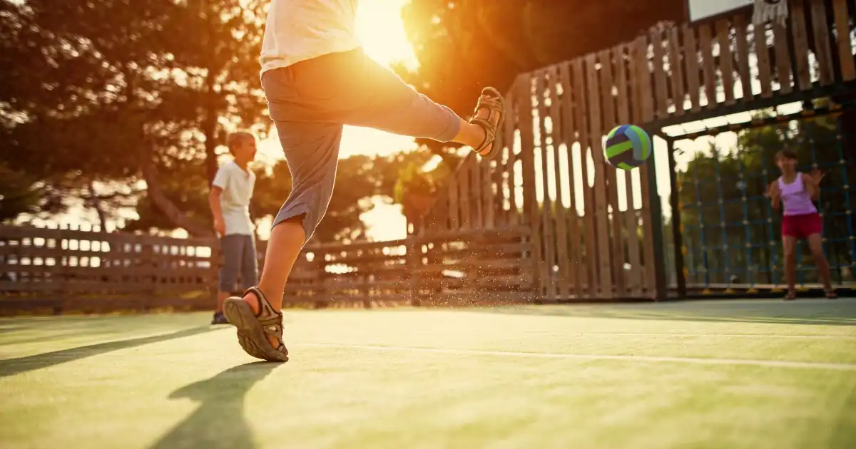 Kind schießt einen Ball auf einem Schulhof-Sportplatz im warmen Abendlicht, ergänzt durch die Aufschrift „Vom behüteten Nest auf den großen Schulhof“.