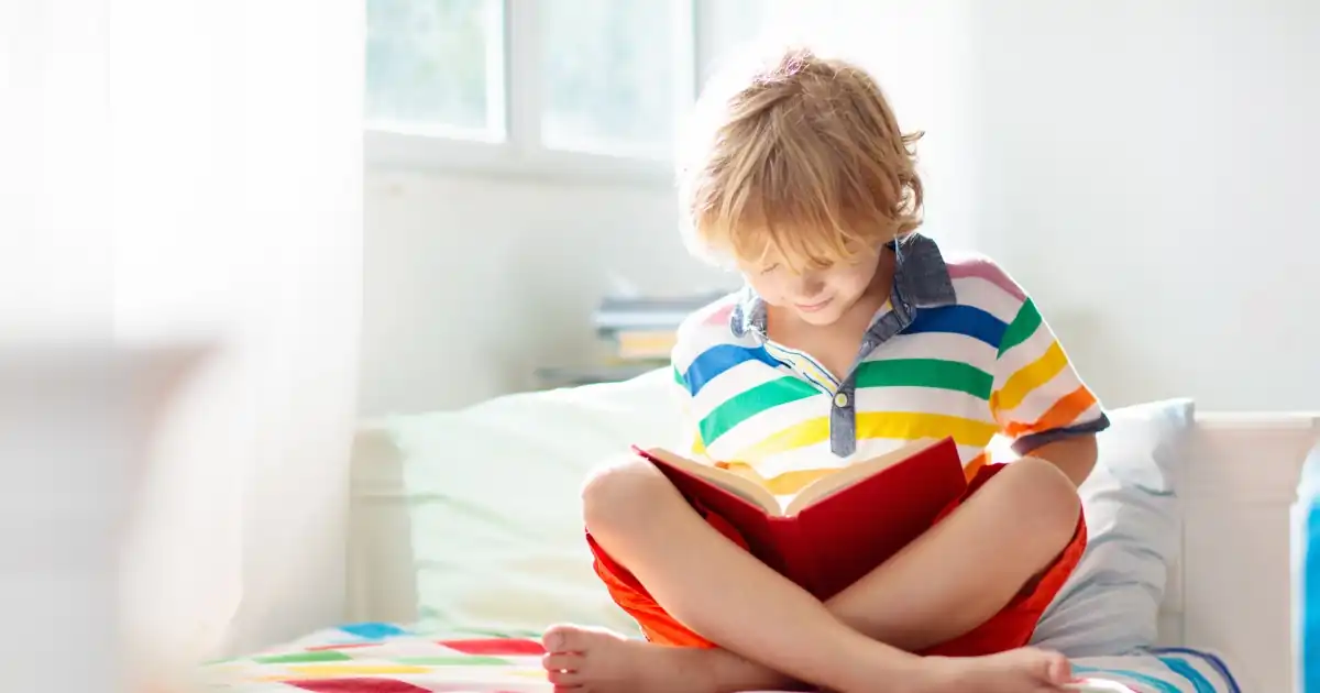 Hell ausgeleuchtetes Kinderzimmer: Ein kleiner Junge mit blonden Haaren und einem bunt gestreiften Poloshirt sitzt barfuß im Schneidersitz auf seinem Bett. Er blickt aufmerksam und konzentriert in ein aufgeschlagenes Buch mit rotem Einband.