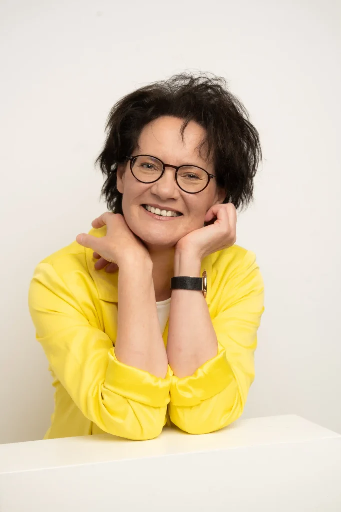 Ein helles Porträtfoto von Petra Bettermann vor einem weißen Hintergrund. Sie stützt die Ellbogen auf eine weiße Fläche, legt die Hände locker an Hals und Kinn und lächelt fröhlich direkt in die Kamera. Sie hat kurze, dunkle, leicht wuschelige Haare und trägt eine dunkle Brille. Auffällig ist ihre leuchtend gelbe Jacke mit hochgekrempelten Ärmeln, unter der ein weißes Oberteil hervorschaut. An ihrem linken Handgelenk ist eine dunkle Armbanduhr zu erkennen.