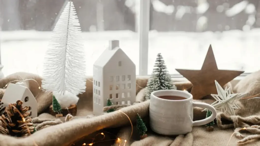 Eine gemütliche winterliche Szene auf einer Fensterbank: Im Vordergrund steht eine weiße Keramiktasse mit einem Heißgetränk auf einer weichen, beigefarbenen Wolldecke, durchflochten von einer warm leuchtenden Lichterkette. Dazwischen sind weiße Keramikhäuschen, kleine grüne und weiße Tannenbäume, ein Tannenzapfen sowie Holz- und Dekosterne arrangiert. Durch das Fenster im Hintergrund blickt man auf eine unscharfe, verschneite Winterlandschaft.