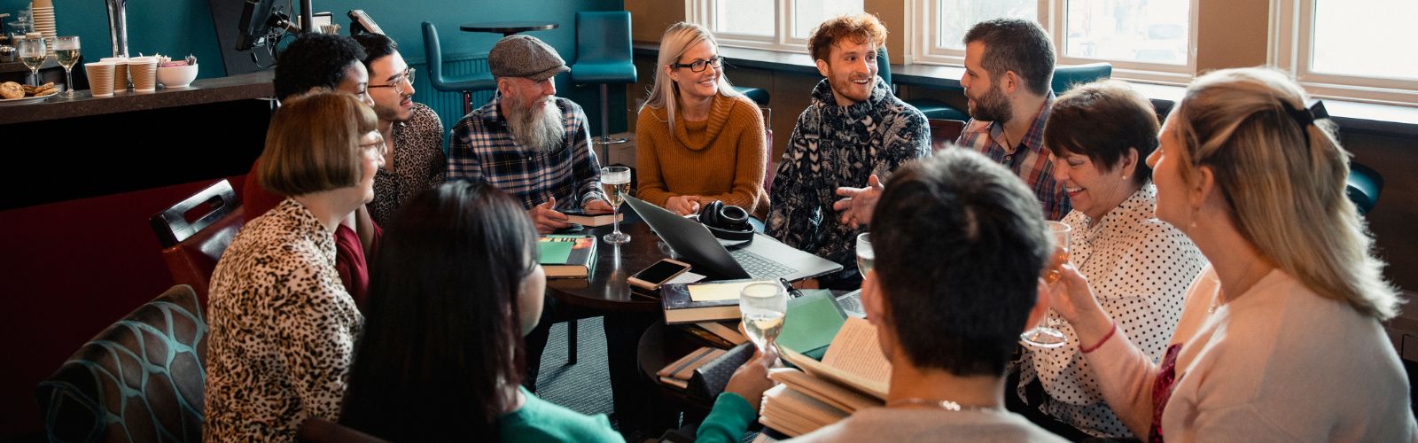 Eine Gruppe von Menschen unterschiedlichen Alters und Stils sitzt in einem gemütlichen Café an einem runden Tisch zusammen. Auf dem Tisch liegen mehrere aufgeschlagene Bücher, Laptops, Notizhefte und Gläser mit Getränken. Die Teilnehmer unterhalten sich lebhaft und lächelnd, gestikulieren und hören einander aufmerksam zu – ganz im Stil eines Treffens eines Buchclubs oder einer Diskussionsrunde. Im Hintergrund ist ein Bar-Bereich sowie Tageslicht durch große Fenster zu sehen.  