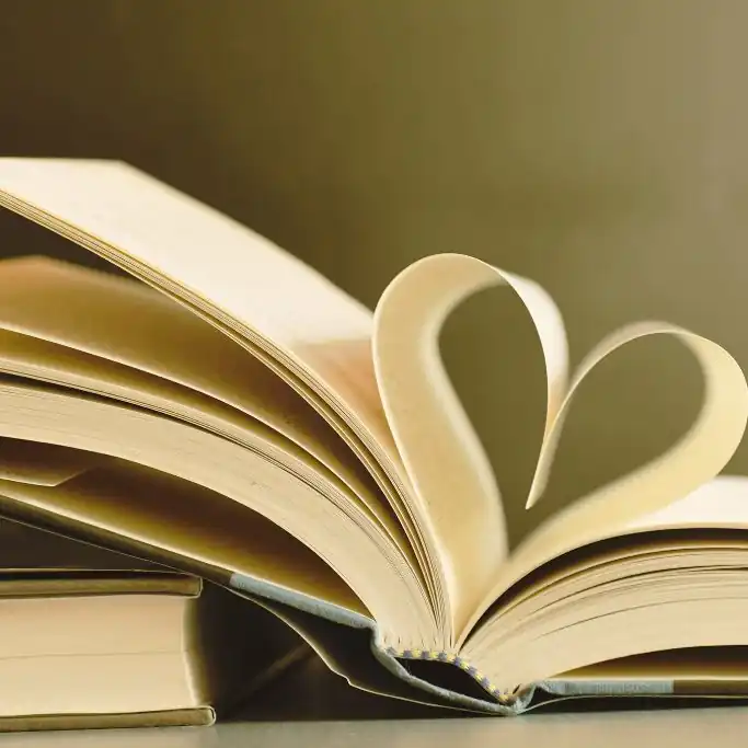 Eine Detailaufnahme eines geöffneten Buches, dessen mittlere Seiten zur Buchmitte hin so geschwungen gefaltet sind, dass sie die Form eines Herzens bilden. Das Buch liegt auf einem weiteren geschlossenen Buch vor einem weichgezeichneten, warmen Hintergrund.
