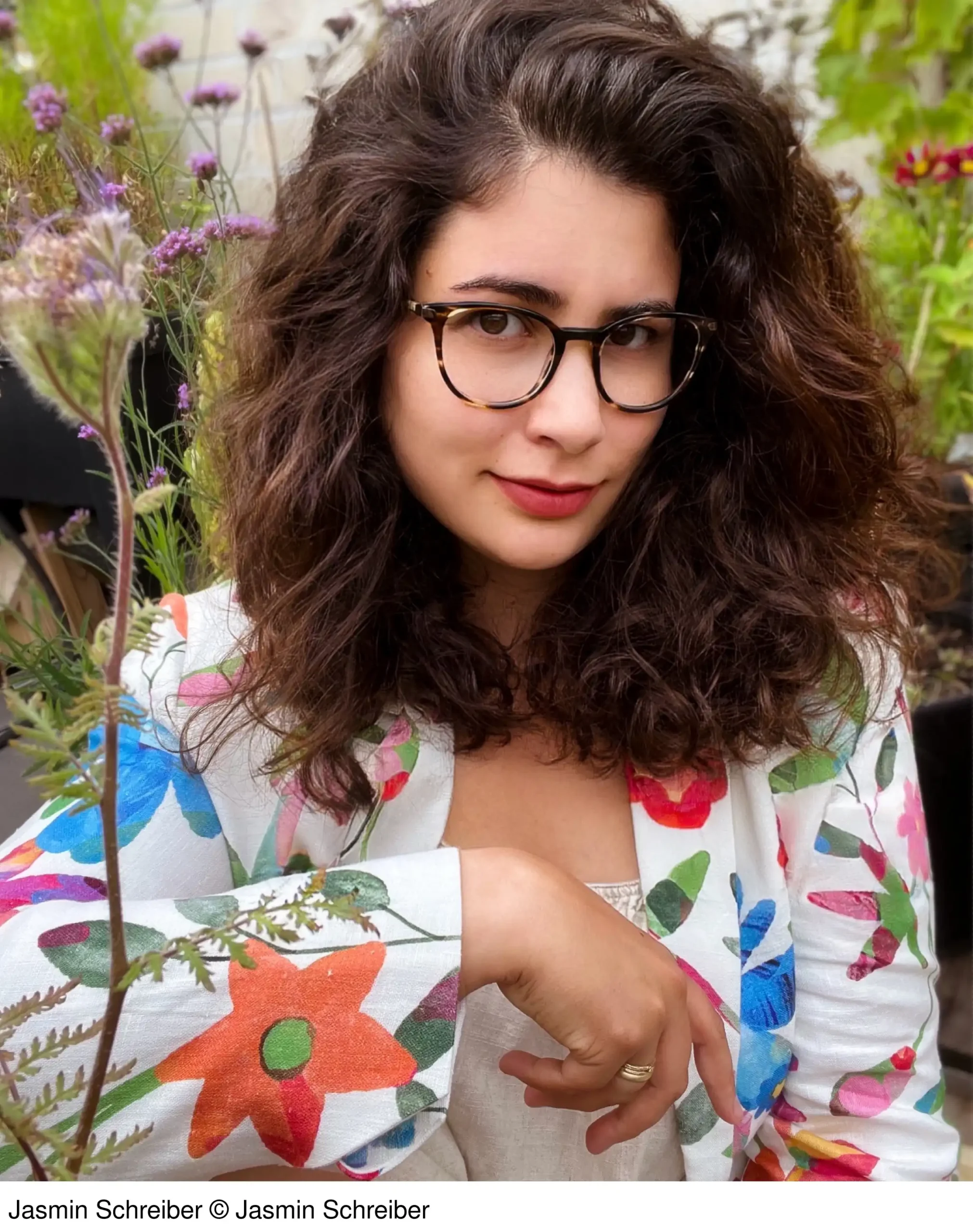 Ein Porträt von Jasmin Schreiber, die mit langen, dunklen Locken und einer Hornbrille direkt in die Kamera lächelt. Sie trägt eine weiße Jacke mit einem auffälligen, bunten floralen Muster. Der Hintergrund ist ein unscharfer Garten mit vielen grünen Pflanzen und lila Blüten. Ihre linke Hand mit einem Ehering ist sichtbar.