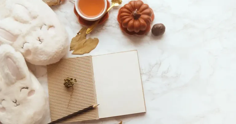 Eine gemütliche Flatlay-Aufnahme auf einer hellen Marmoroberfläche zum Thema Journaling: In der Mitte liegt ein geöffnetes Notizbuch mit leeren Seiten, einem Umschlag aus brauner Wellpappe und einem dunklen Bleistift. Links sind zwei flauschige, weiße Hausschuhe in Hasenoptik zu sehen. Im oberen Bildbereich sorgen eine kleine Tasse Tee mit einem goldenen Löffel, ein terrakottafarbener Dekokürbis, getrocknete Blätter und feine Naturdetails für eine herbstliche Atmosphäre.