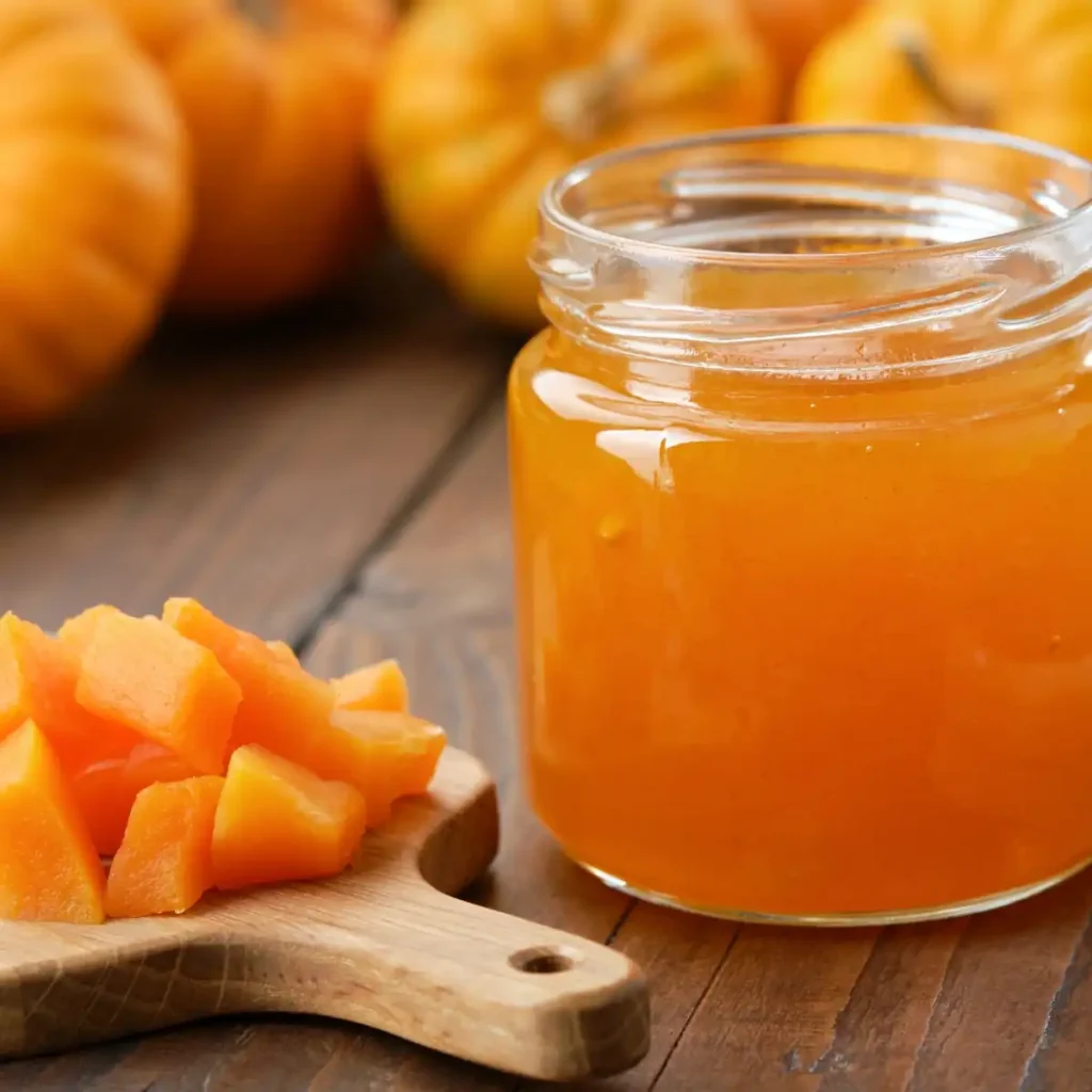 Eine herbstliche Nahaufnahme auf einem hölzernen Untergrund: Rechts steht ein geöffnetes Glas gefüllt mit leuchtend orangener Kürbismarmelade. Links daneben liegen frische, würfelförmig geschnittene Kürbisstücke auf einem kleinen Holzbrettchen. Im weichgezeichneten Hintergrund sind mehrere ganze Kürbisse zu erkennen.