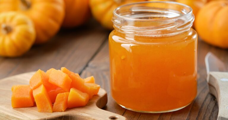 Eine herbstliche Nahaufnahme auf einem hölzernen Untergrund: Rechts steht ein geöffnetes Glas gefüllt mit leuchtend orangener Kürbismarmelade. Links daneben liegen frische, würfelförmig geschnittene Kürbisstücke auf einem kleinen Holzbrettchen. Im weichgezeichneten Hintergrund sind mehrere ganze Kürbisse zu erkennen.