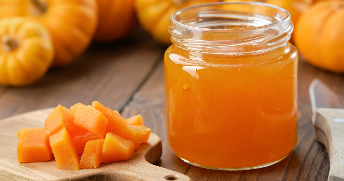Eine herbstliche Nahaufnahme auf einem hölzernen Untergrund: Rechts steht ein geöffnetes Glas gefüllt mit leuchtend orangener Kürbismarmelade. Links daneben liegen frische, würfelförmig geschnittene Kürbisstücke auf einem kleinen Holzbrettchen. Im weichgezeichneten Hintergrund sind mehrere ganze Kürbisse zu erkennen.