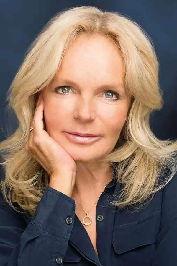 Ein Porträtfoto der Autorin Lucinda Riley vor einem dunkelblauen Hintergrund. Sie blickt mit einem sanften, geschlossenen Lächeln direkt in die Kamera und stützt ihr Kinn nachdenklich auf ihre rechte Hand. Ihr blondes, schulterlanges Haar umrahmt ihr Gesicht. Sie trägt eine dunkelblaue Bluse, die farblich mit dem Hintergrund harmoniert, und eine feine goldene Halskette mit einem kreisförmigen Anhänger. An ihrer rechten Hand blitzt ein Ring auf. Das Licht hebt ihre Gesichtszüge und ihre hellblauen Augen sanft hervor.