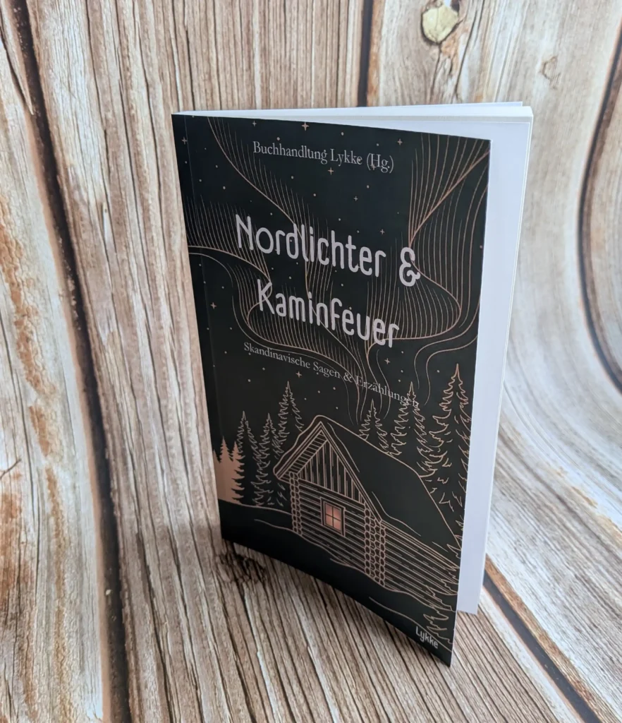 Ein aufrecht auf einer hellen Holzoberfläche stehendes Buch mit dunkelbraunem bis schwarzem Cover. Das Cover zeigt oben den Herausgeber „Buchhandlung Lykke (Hg.)“, den Titel „Nordlichter & Kaminfeuer“ sowie den Untertitel „Skandinavische Sagen & Erzählungen“. Die filigrane, kupferfarben gezeichnete Illustration stellt eine gemütliche Blockhütte mit beleuchtetem Fenster inmitten von Tannenbäumen dar, über der sich geschwungene Polarlichter an einem Sternenhimmel abzeichnen. Unten rechts ist das Verlagslogo „Lykke“ platziert.