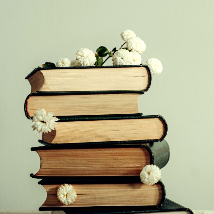 Ein Stapel alter, gebundener Bücher mit dunklen Einbänden vor einem hellen, dezenten Hintergrund. Auf dem obersten Buch liegen kleine, weiße Blüten, während weitere vereinzelte Blütenköpfe zwischen den einzelnen Buchseiten des Stapels stecken.