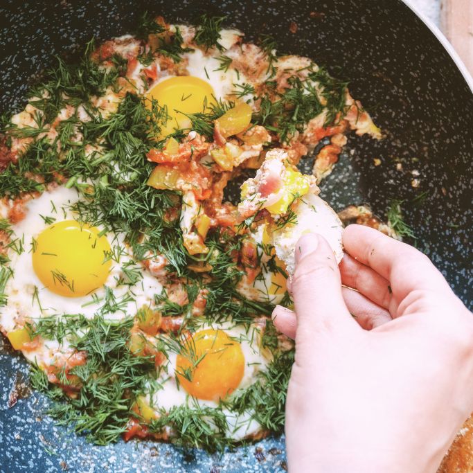 Eine Draufsicht auf eine dunkle Pfanne mit einem herzhaften Eiergericht in Tomaten-Gemüse-Soße (Shakshuka), das reichlich mit frischem, gehacktem Dill bestreut ist. Unten rechts tunkt eine Hand ein Stück weißes Brot in die Pfanne, um etwas von der Speise aufzunehmen.