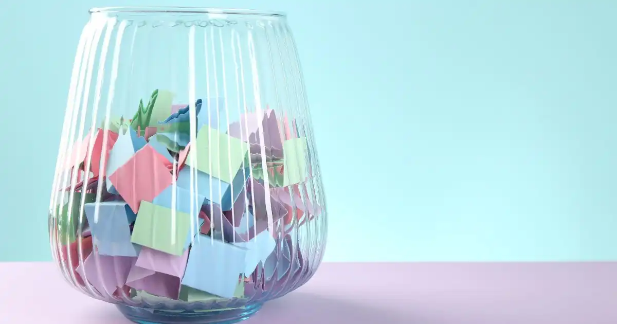 Ein transparenter Glasbehälter auf fliederfarbenem Untergrund, gefüllt mit vielen bunt gefalteten Papierlosen für eine Verlosung vor hellblauem Hintergrund.