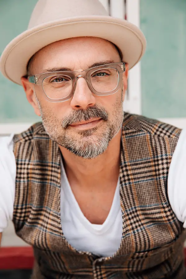 Ein nahes Porträtfoto von Volker Klüpfel, der mit einem ruhigen, leicht angedeuteten Lächeln direkt in die Kamera blickt. Er trägt einen beigefarbenen Filzhut, eine markante Brille mit transparentem Kunststoffgestell und hat einen gepflegten, leicht grau melierten Dreitagebart. Gekleidet ist er in ein weißes T-Shirt und eine ärmellose Weste mit einem klassischen Karomuster in Braun- und Beigetönen. Der Hintergrund ist leicht unscharf und in hellen, pastelligen Grün- und Weißtönen gehalten.